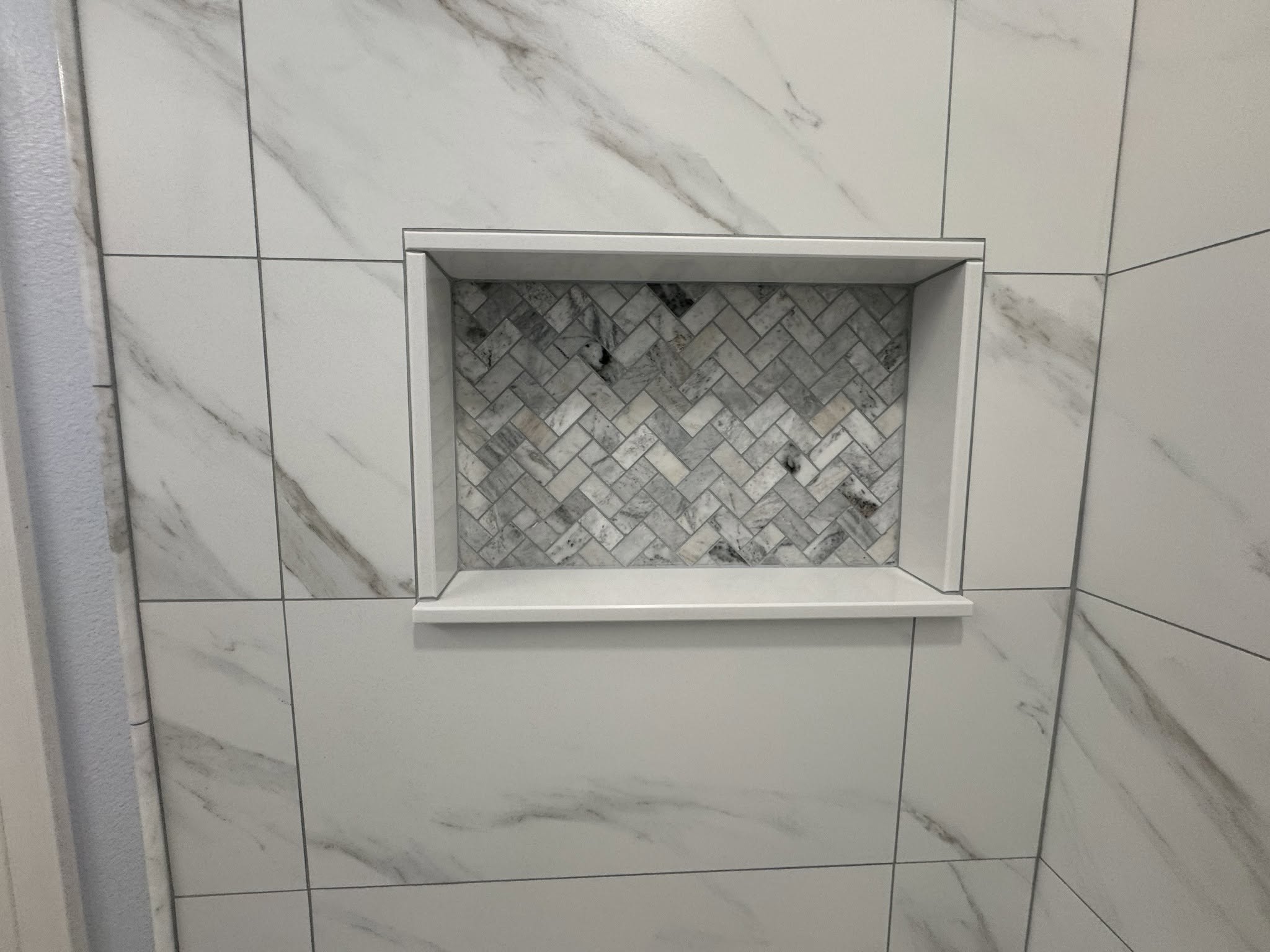 Hand-built shower niche with a herringbone mosaic back, white border and shelf, set into marble-look wall tile.