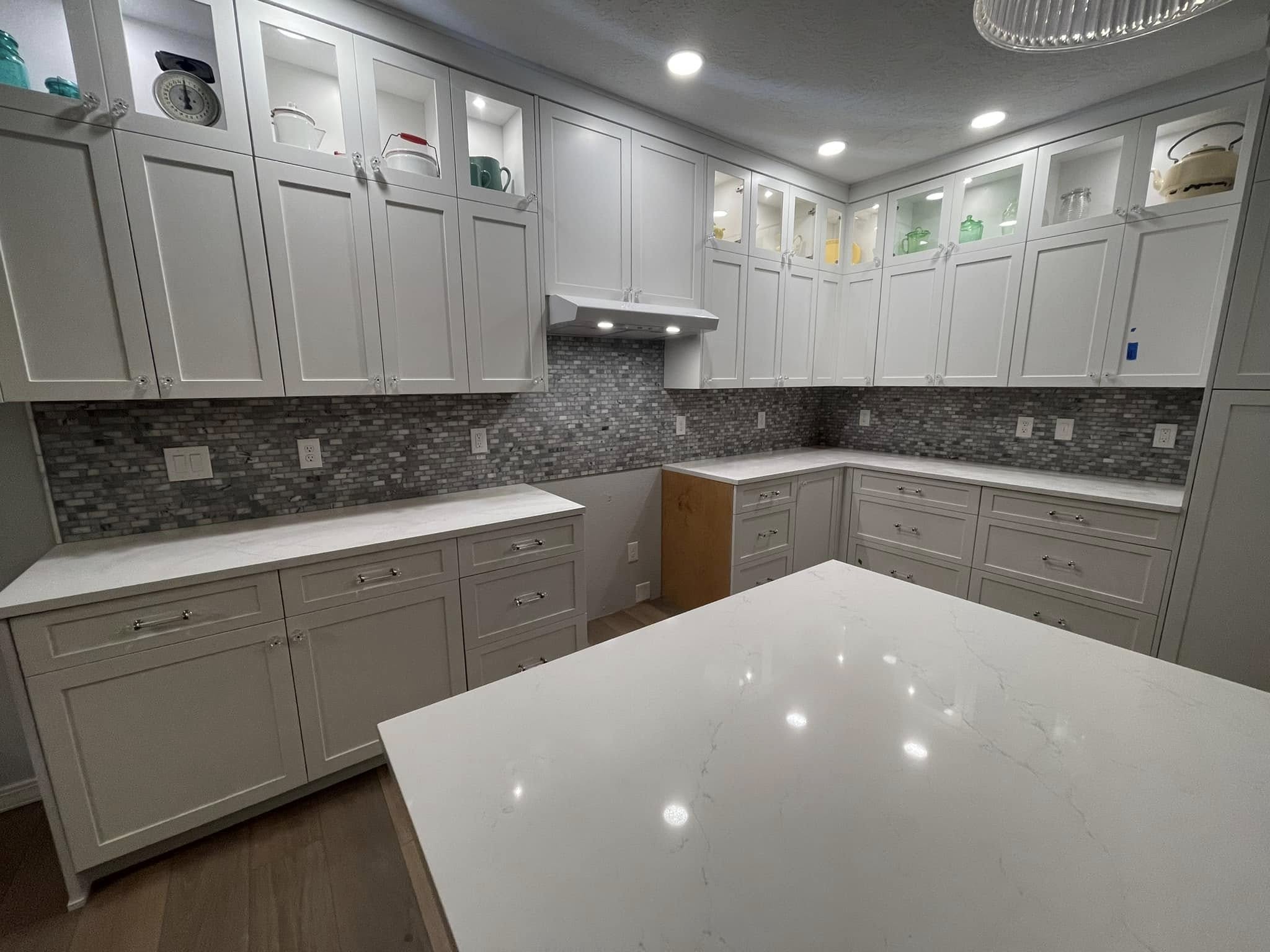White kitchen cabinetry with illuminated display cupboards, mosaic backsplash and a pale countertop. Photographed before final appliance installation.